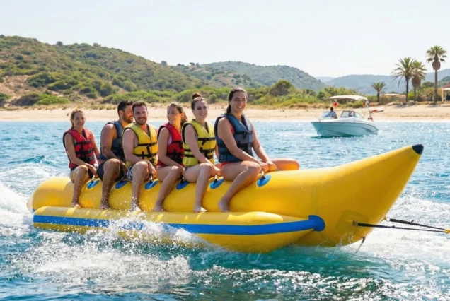 A group of six people wearing life vests are enjoying a ride on a yellow banana boat in the ocean near a sandy beach with hills and palm trees.