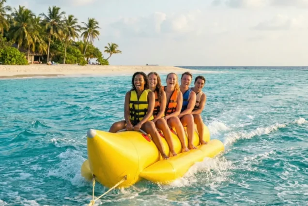 Five people in life jackets smiling on a yellow banana boat ride in tropical blue waters, near a beach with palm trees under a cloudy sky. Fun and adventure.