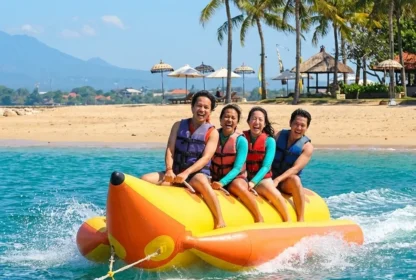 Four people in life vests are joyfully riding a banana boat on turquoise waters near a sandy beach with palm trees and distant mountains.