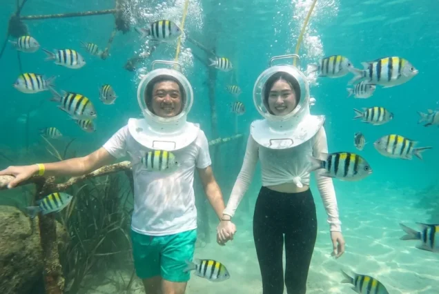 A person wearing a white oxygen helmet walking on the ocean floor in Nusa Dua surrounded by coral.