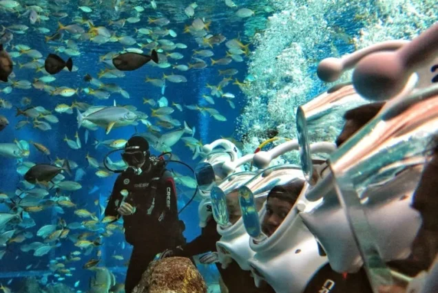 A tourist wearing a white oxygen helmet walking on the ocean floor in Nusa Dua surrounded by tropical fish and coral reefs.