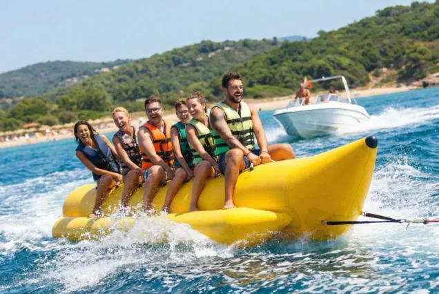 A group of six people wearing colorful life vests enjoy a ride on a yellow banana boat on clear blue water, with a speedboat in the background.