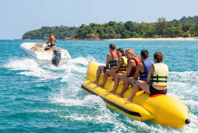 Five people on a yellow banana boat are towed by a speedboat on clear blue water near a sandy beach, exuding joy and adventure.