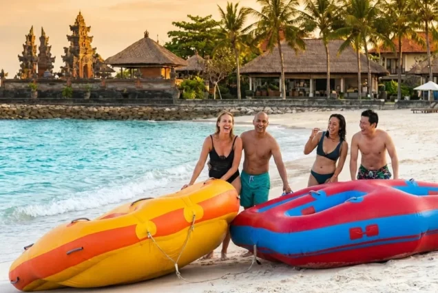Four people laughing on a sandy beach with colorful inflatable rafts. Palm trees and traditional architecture in the background evoke a tropical vibe.