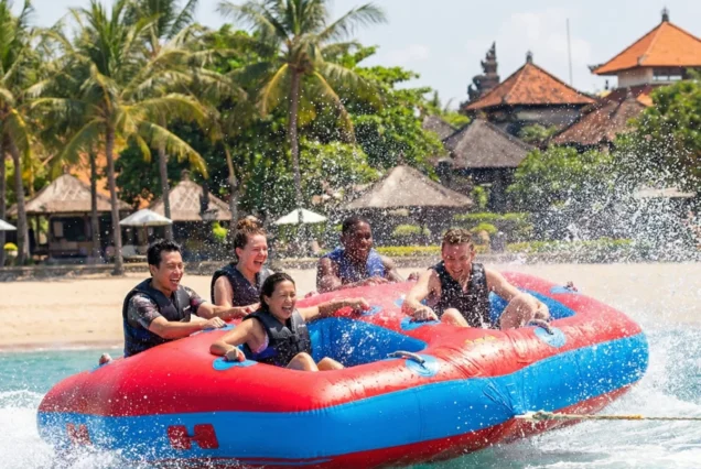 Five people wearing life vests enjoy a thrilling ride on an inflatable raft, with water splashing around. Palm trees and beach huts are visible in the background.