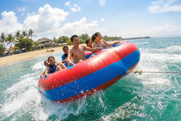A group enjoys a thrilling ride on a red and blue inflatable raft on clear, blue ocean waters. Waves splash around them under a sunny, tropical sky.