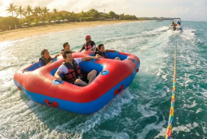 A group of five people enjoy tubing on a large red and blue inflatable raft, being towed by a boat. They laugh and smile, surrounded by tropical blue water with a sandy beach and palm trees in the background under a bright, sunny sky.
