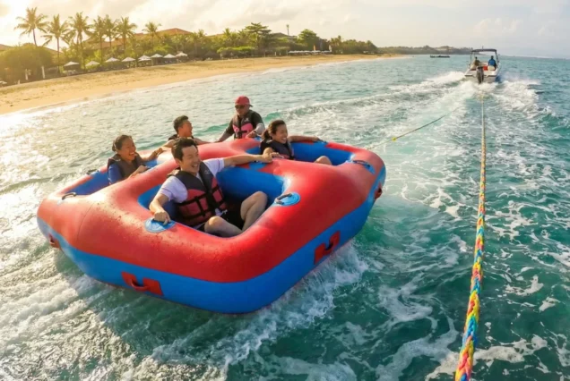 A group of five people enjoy tubing on a large red and blue inflatable raft, being towed by a boat. They laugh and smile, surrounded by tropical blue water with a sandy beach and palm trees in the background under a bright, sunny sky.