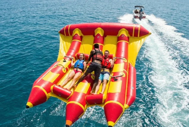 Three people in colorful life jackets enjoy an inflatable ride, towed by a boat on clear blue water. Vibrant and joyful atmosphere.