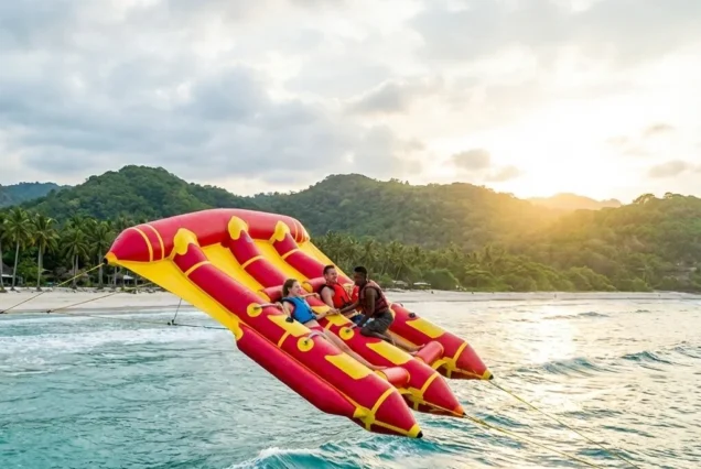 Three people enjoy a thrilling ride on a red and yellow inflatable raft pulled across blue ocean waters, with a sunset and palm-lined shore in the background.