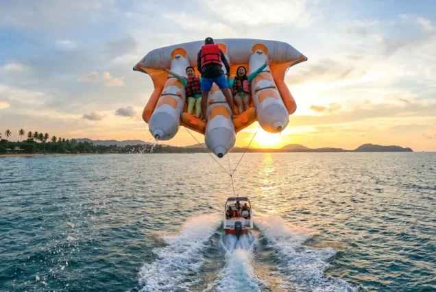 Three people in life jackets enjoy an inflatable ride over the ocean at sunset, towed by a speedboat. The mood is adventurous and joyful.