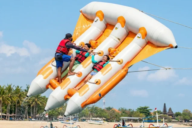 Three people in life vests enjoy an orange inflatable ride, skimming above the sea, with a clear blue sky and palm trees in the background.