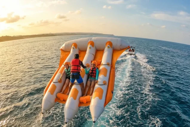 A bright, orange inflatable water raft, towed by a boat, hovers above the ocean with three excited people wearing life jackets, against a sunset sky.