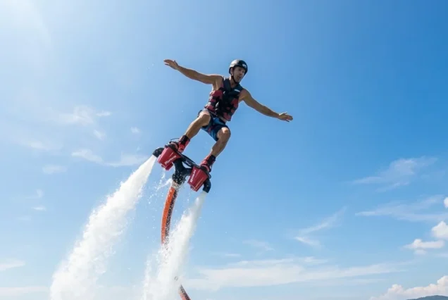 A person wearing a helmet and life vest flyboards over water, propelled by powerful jets. The sky is clear blue, evoking a sense of freedom and adventure.
