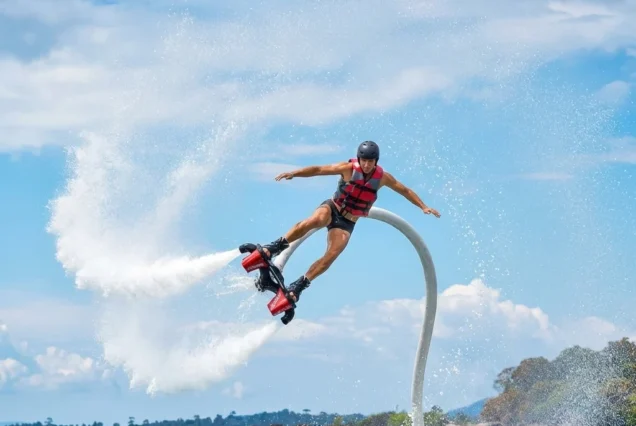 A person wearing a helmet and life vest is flyboarding over the ocean, performing an aerial maneuver, with water spraying around them under a clear blue sky.