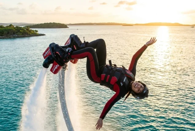 A person joyfully rides a water-powered flyboard, performing a flip against a sunset backdrop over the ocean. They wear a black and red wetsuit and helmet.