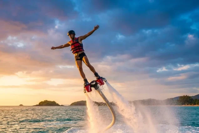 A man in a red life jacket flyboarding over the ocean at sunset, with arms outstretched. The sky is partly cloudy, with orange and blue hues.