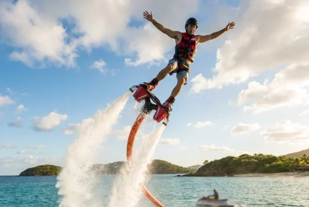 A person joyfully flyboarding above clear ocean water, arms outstretched. The backdrop is a sunny sky with clouds and a distant island, conveying excitement.